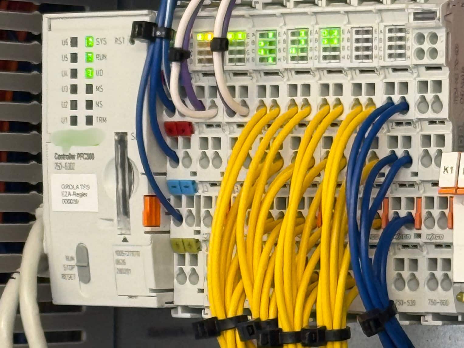 Innenansicht eines Schaltschranks mit einem EZA-Regler. Grün leuchtende Status-LEDs zeigen den Betrieb an. Zahlreiche gelbe und blaue Kabel sind mit Kabelbindern gebündelt und an Klemmenmodulen angeschlossen.